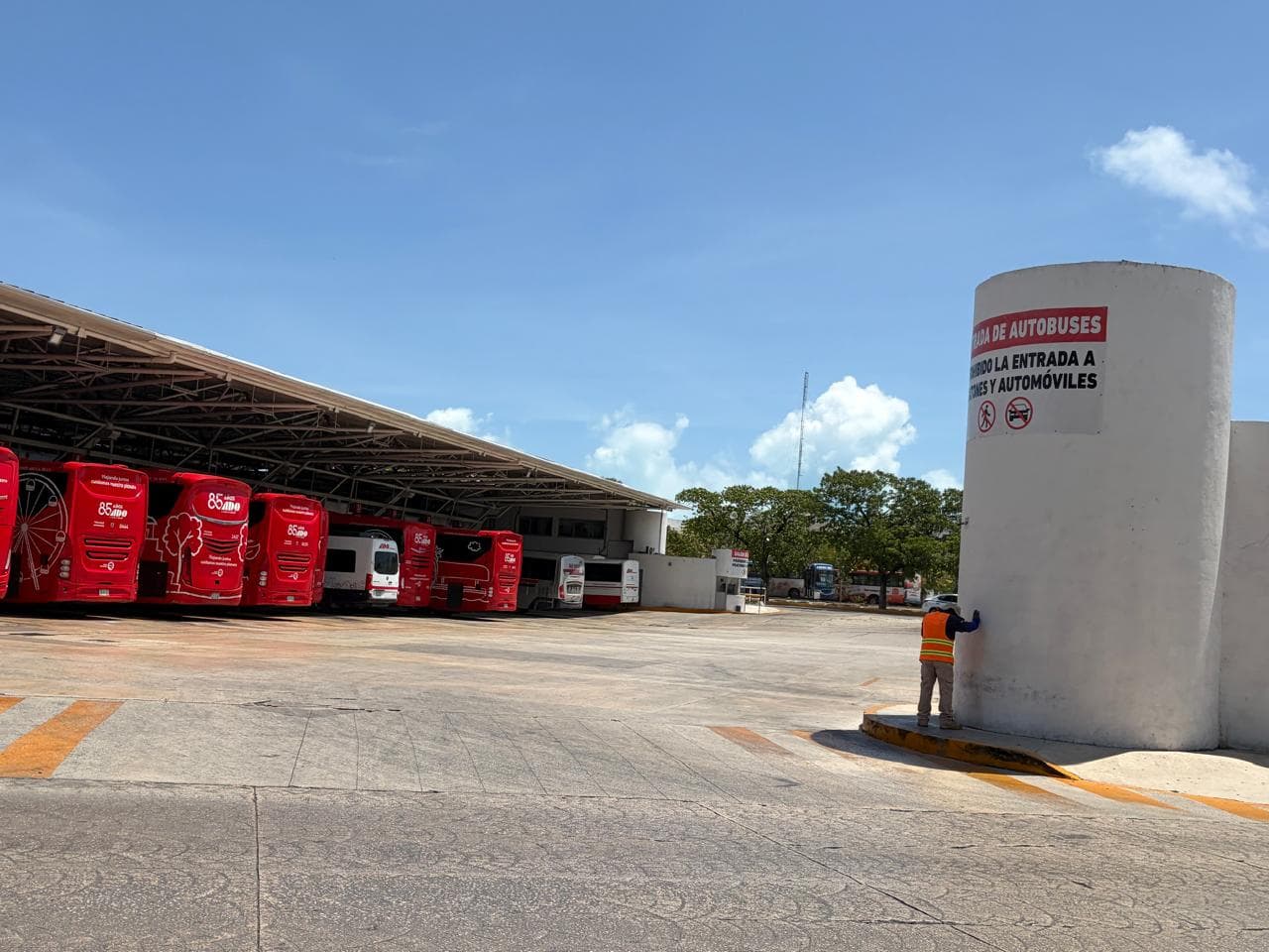 Autobuses rojos de ADO estacionados en la terminal, con un trabajador realizando labores de mantenimiento junto a un tanque de agua.