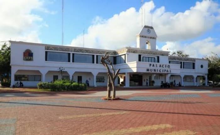 Fachada frontal del Palacio Municipal de Playa del Carmen, edificio de dos pisos con arquitectura colonial, torre del reloj y plaza pavimentada.