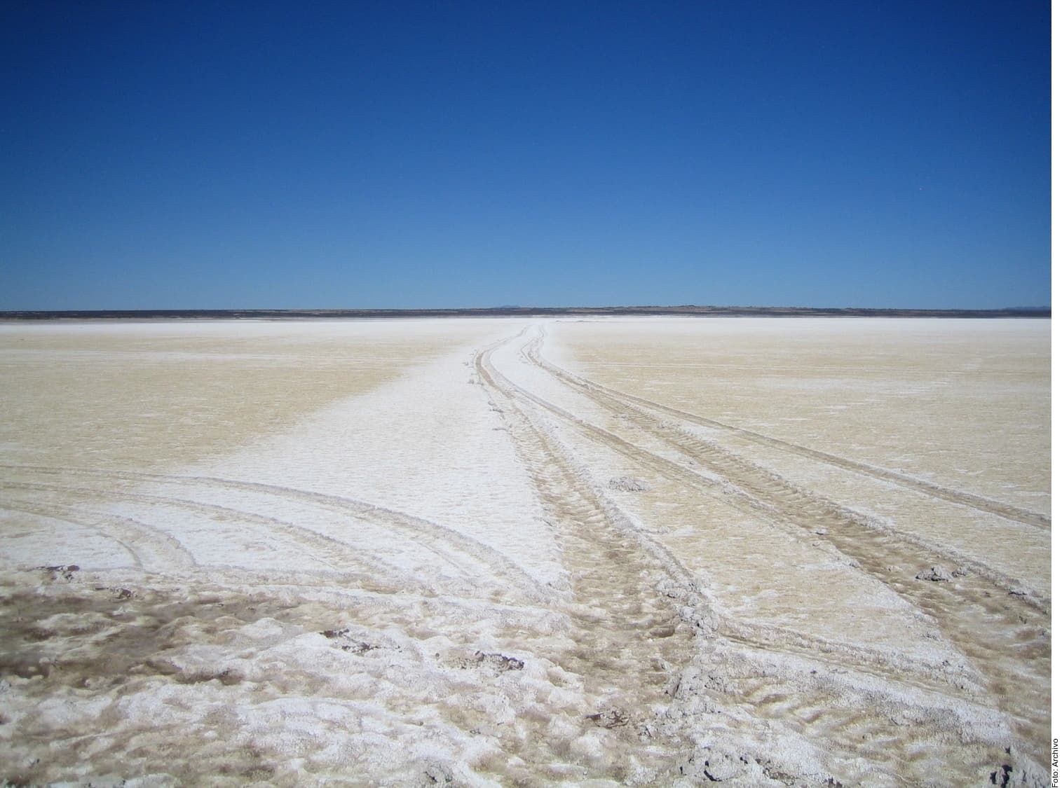 Carretera de tierra sinuosa atravesando un extenso terreno desértico de tonos blancos y beige bajo un cielo azul despejado.