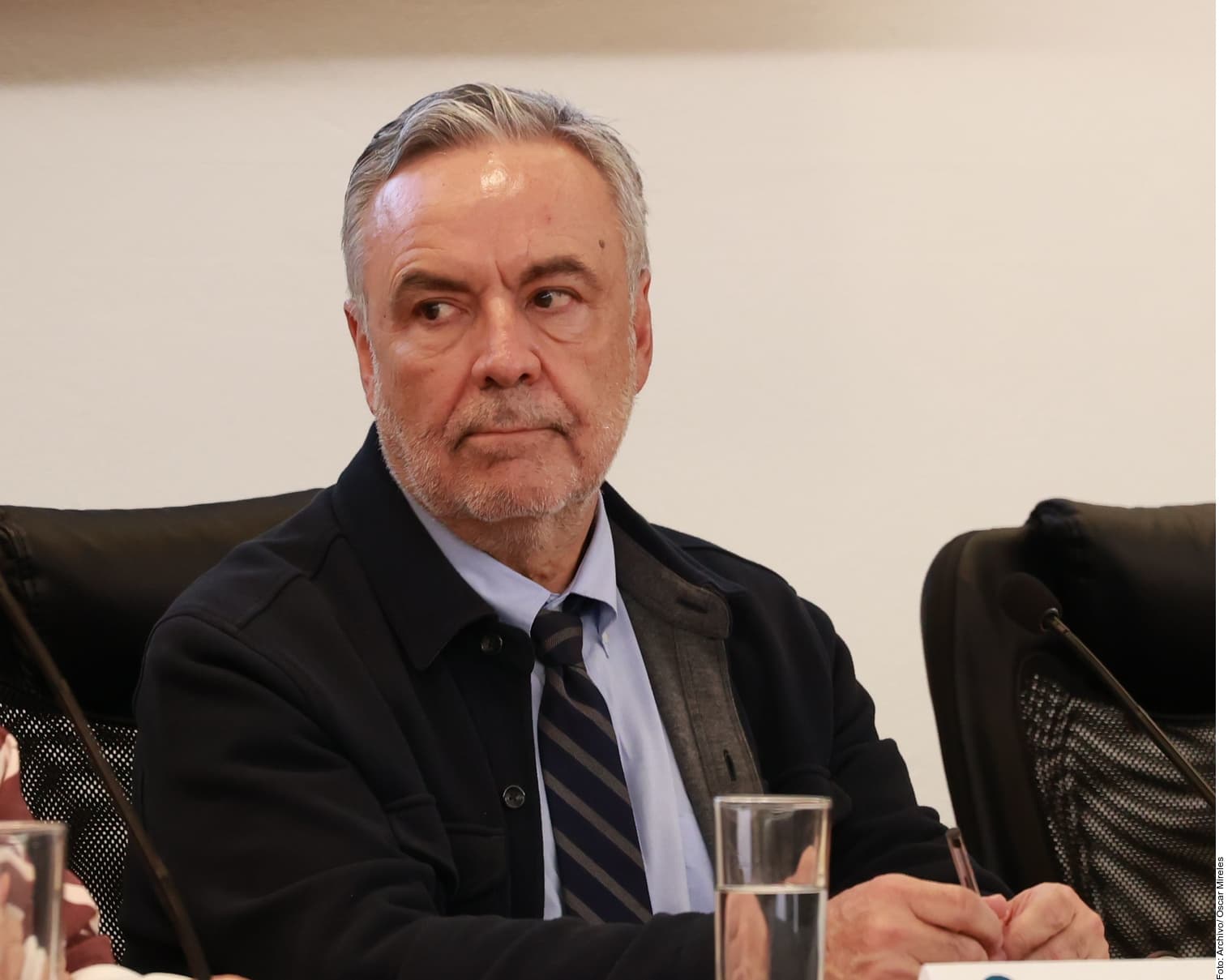 Hombre de traje y corbata en reunión formal sentado ante micrófono y vaso de agua.