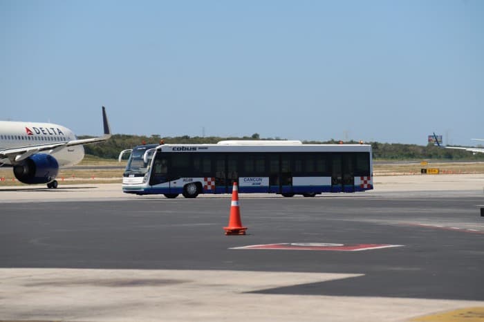 Autobús de transporte de pasajeros circulando en la pista del aeropuerto de Cancún junto a una aeronave Delta estacionada.