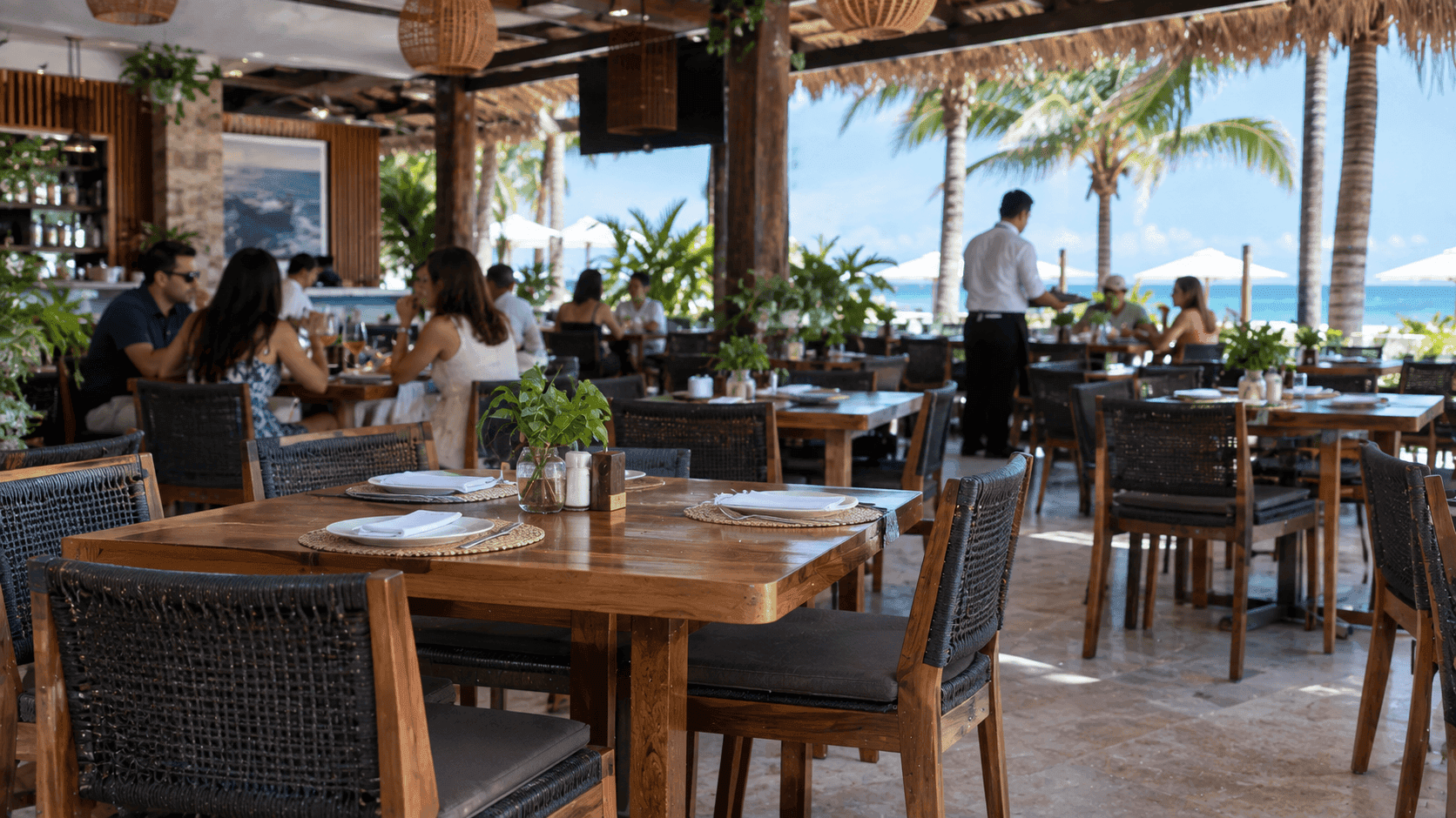 Interior de restaurante de lujo frente al mar con mesas de madera, sillas tejidas, plantas decorativas y clientes disfrutando en ambiente tropical.