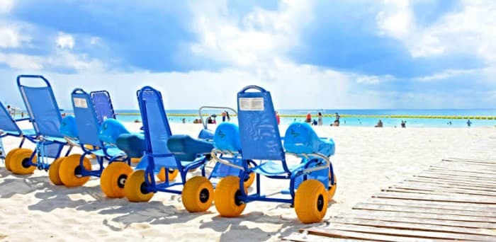 Sillas de ruedas anfibias azules con ruedas amarillas estacionadas en la arena de una playa con turistas al fondo.