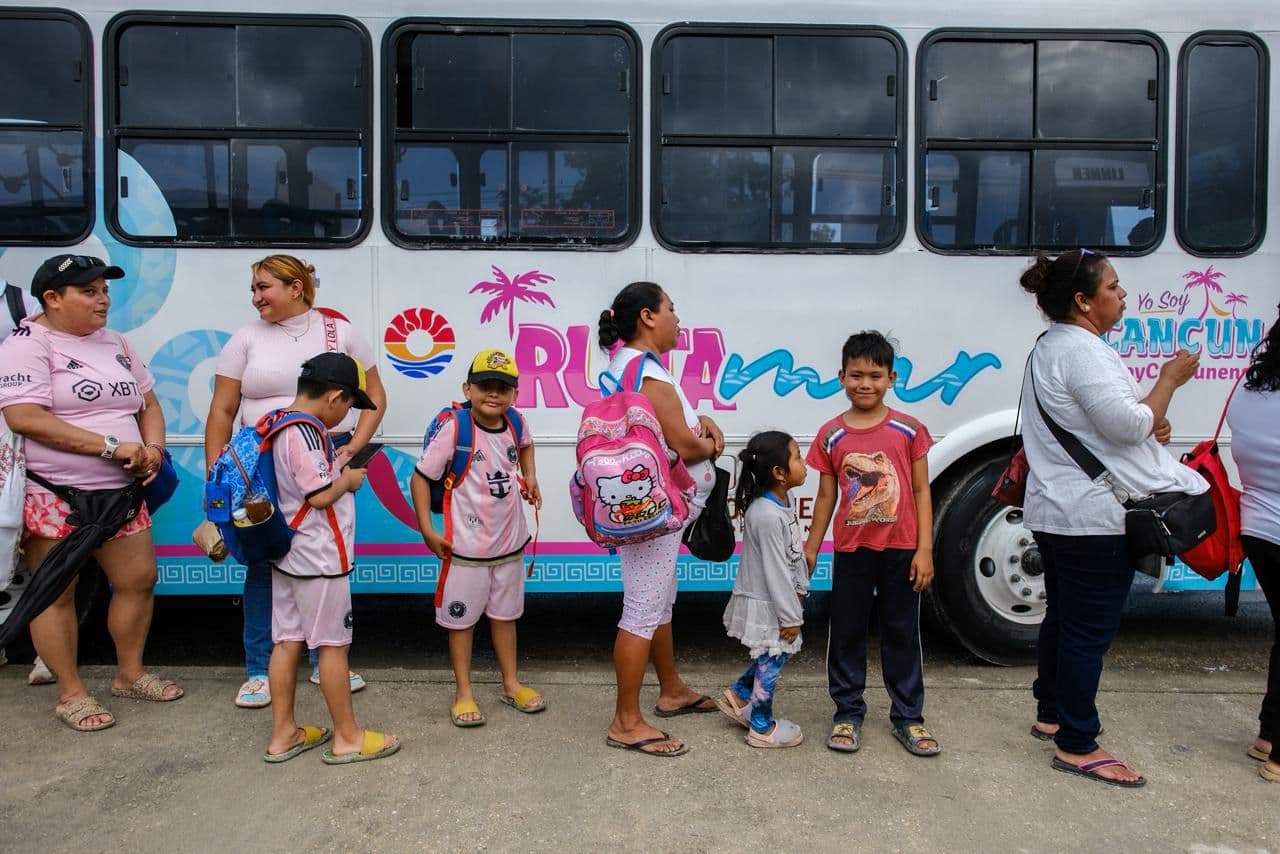 Grupo de residentes y niños esperan junto a un autobús blanco con diseño turístico en Cancún.