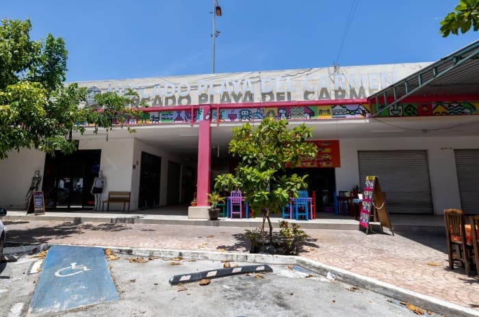Fachada frontal del Mercado Playa del Carmen con entrada principal, columna rosa, señalización y estacionamiento, bajo cielo despejado.