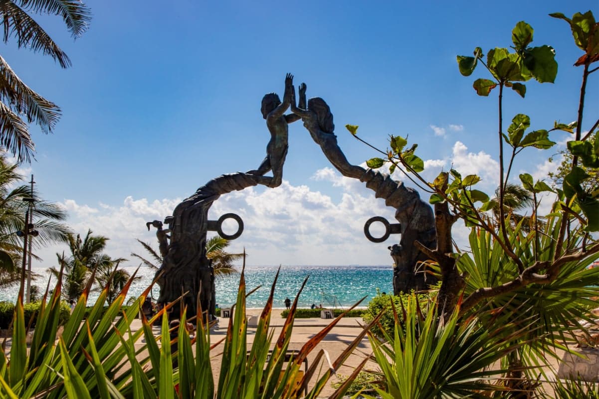 Escultura artística de dos figuras en la playa de Playa del Carmen con palmeras, arena y mar turqueso de fondo.