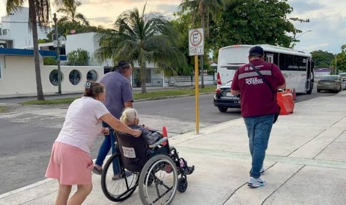 Persona en silla de ruedas siendo asistida por dos personas en una parada de autobús en Cancún, con un vehículo de transporte de fondo.