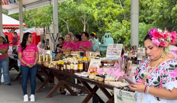 Vendedoras en puesto de productos artesanales durante evento comunitario bajo estructura techada con árboles de fondo.