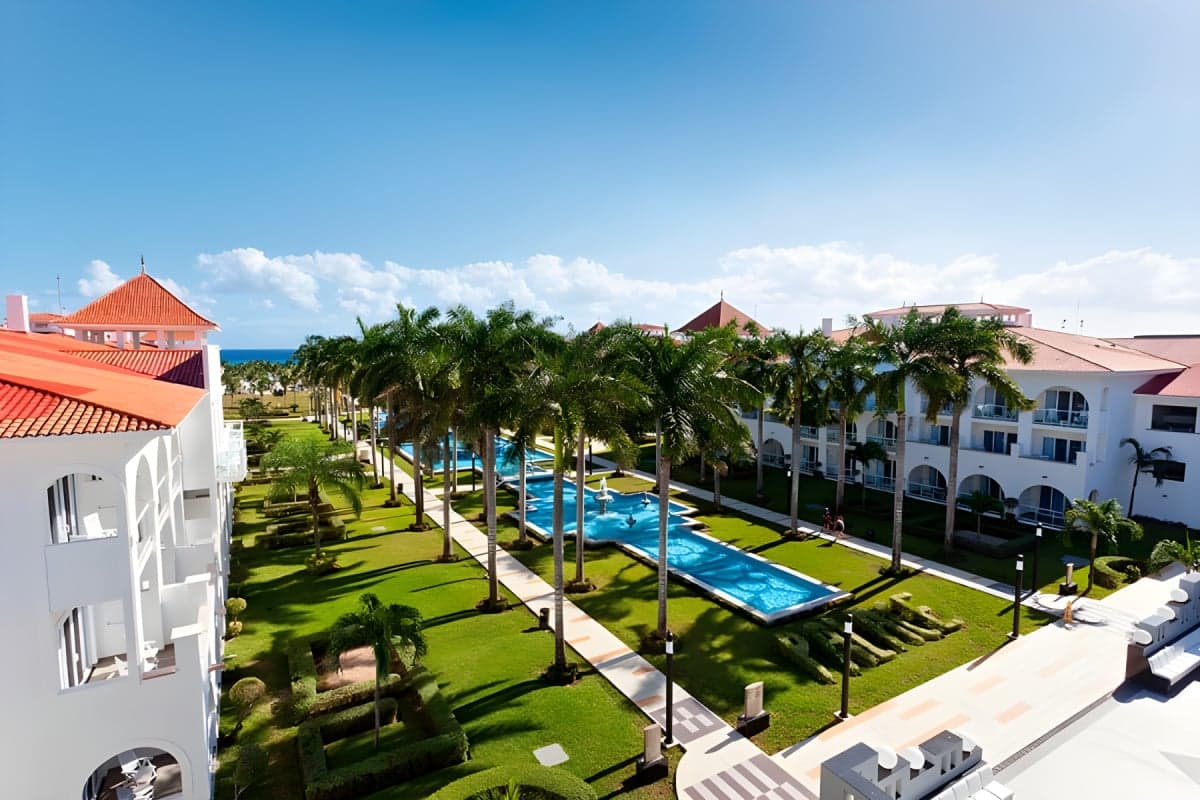 Vista aérea del complejo hotelero Riu Palace Mexico en Playacar con áreas verdes, piscinas azules, edificios blancos con techos de tejas rojas y palmeras tropicales frente al mar Caribe.