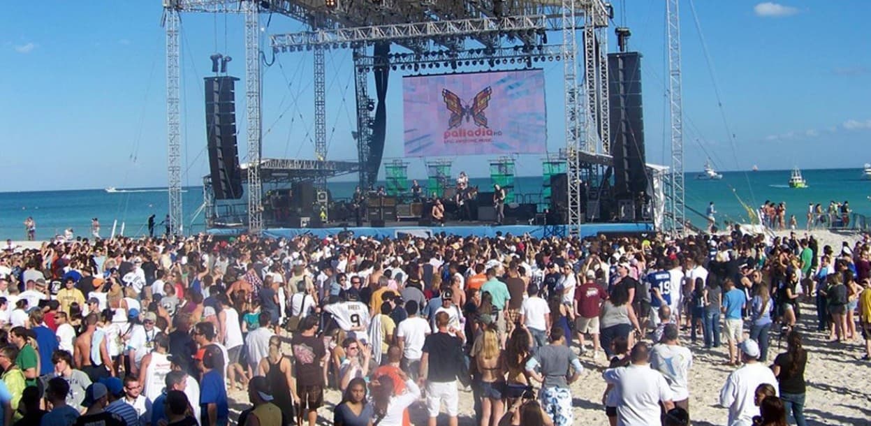 Multitud de turistas en playa de arena blanca frente a escenario con pantalla gigante durante evento musical en la Riviera Maya, con mar turqueso y cielo despejado de fondo.