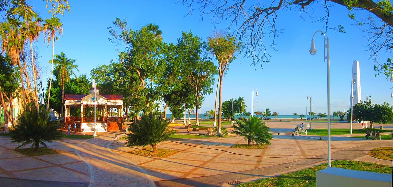 Plaza pública frente al mar con áreas verdes, malecón, palmeras y estructuras turísticas en Quintana Roo bajo cielo despejado.