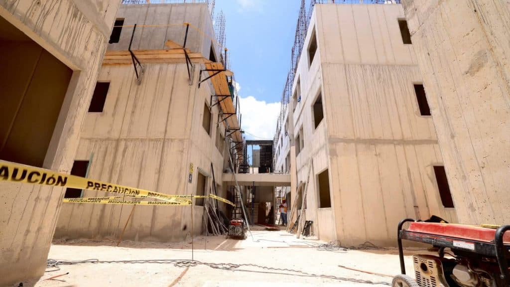 Obra en construcción con edificios de concreto en proceso de desarrollo, cinta de precaución amarilla y equipo de trabajo en el sitio.