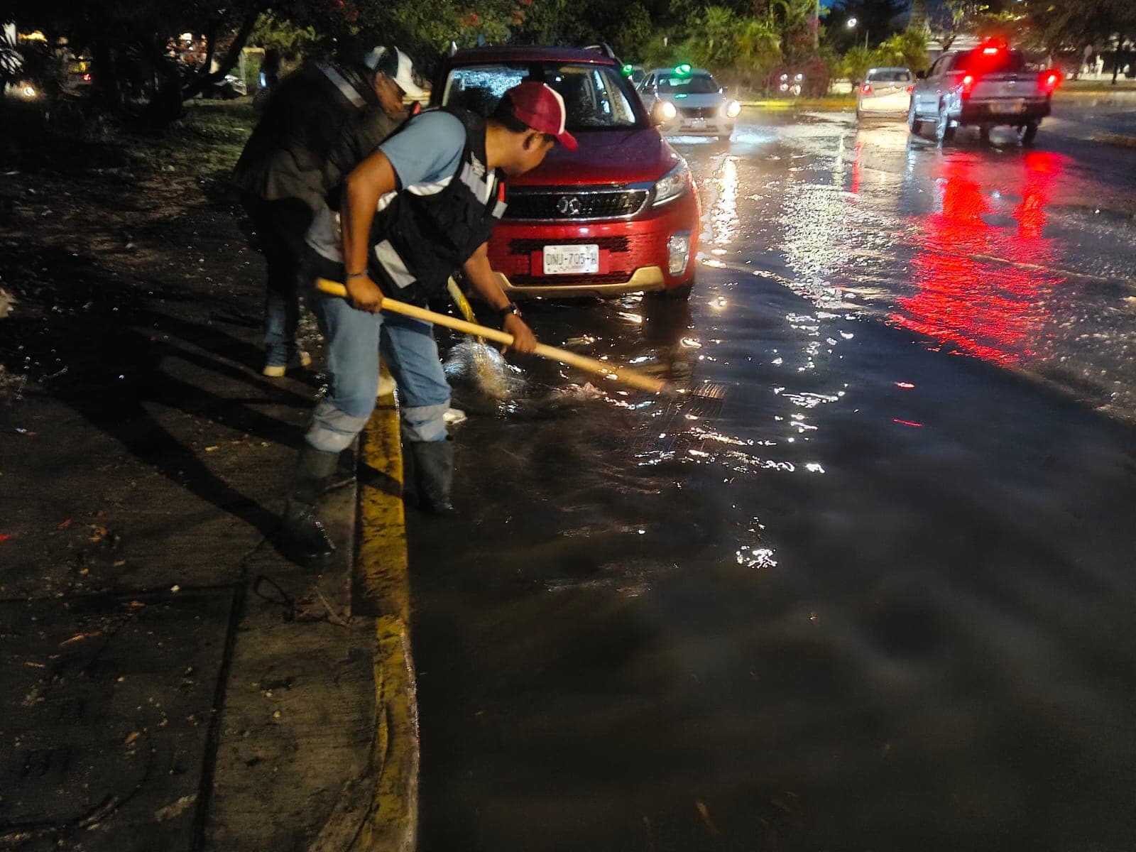 Dos trabajadores con herramientas realizan desazolve en una calle inundada de Playa del Carmen durante la noche, con vehículos y semáforos de fondo.