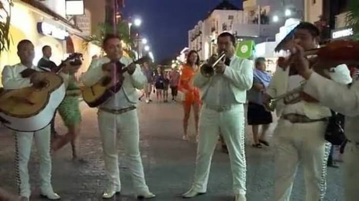 Grupo de mariachis vestidos de blanco tocan instrumentos en una calle peatonal nocturna de una zona comercial.