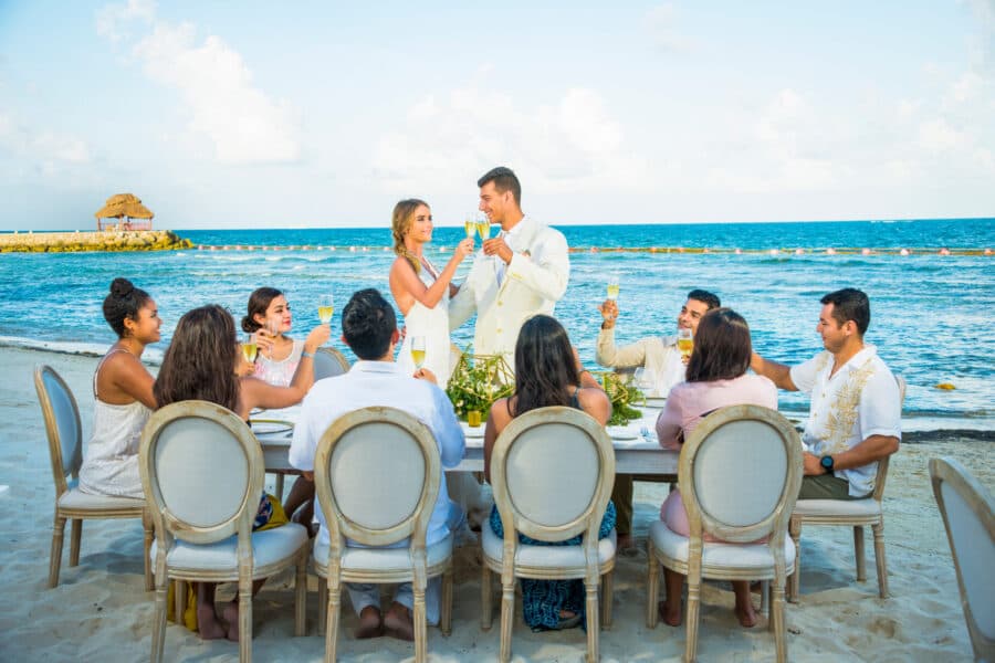 Pareja de novios brinda con invitados durante una ceremonia de boda en la playa de la Riviera Maya con el mar de fondo.