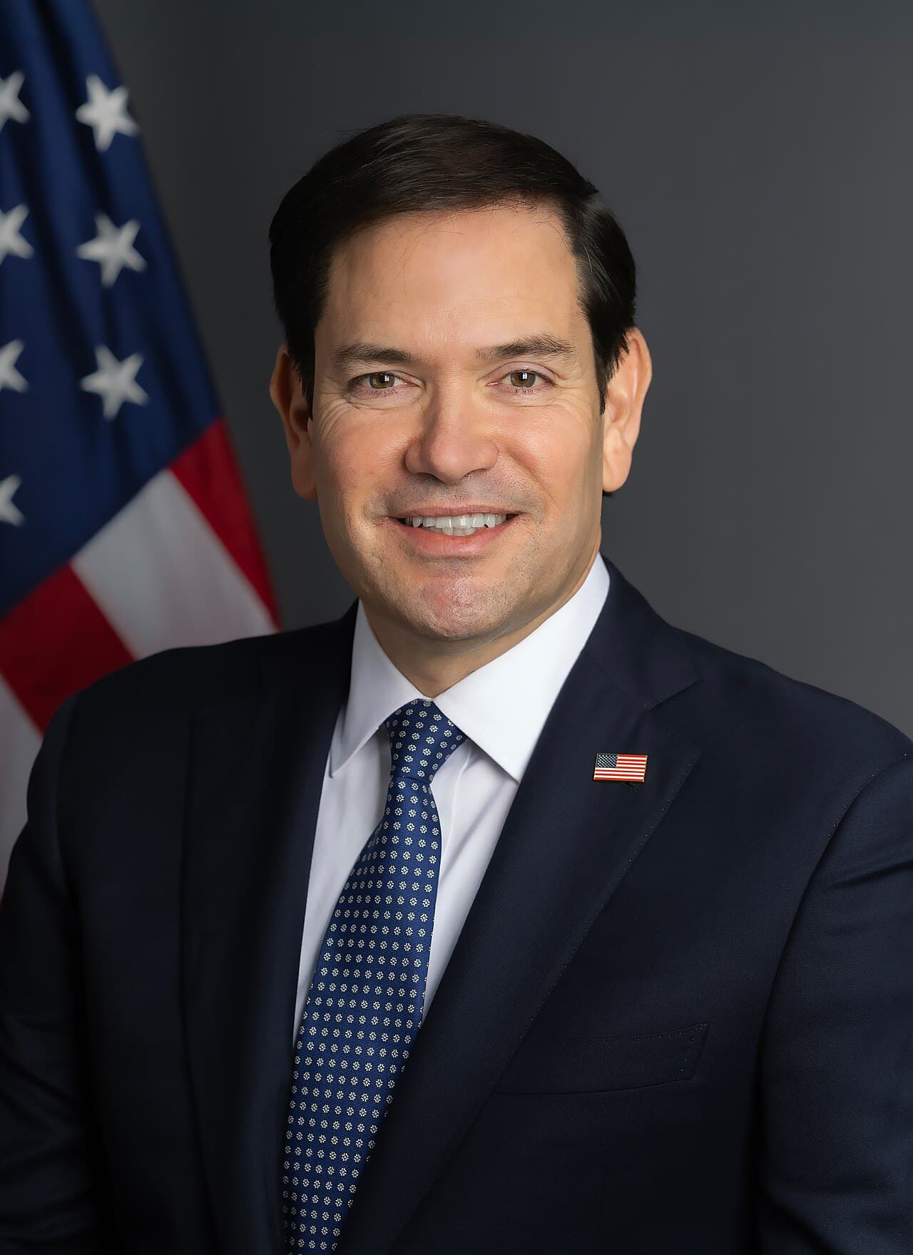 Retrato oficial de un funcionario estadounidense frente a bandera americana, vistiendo traje oscuro y corbata azul con pin de bandera.