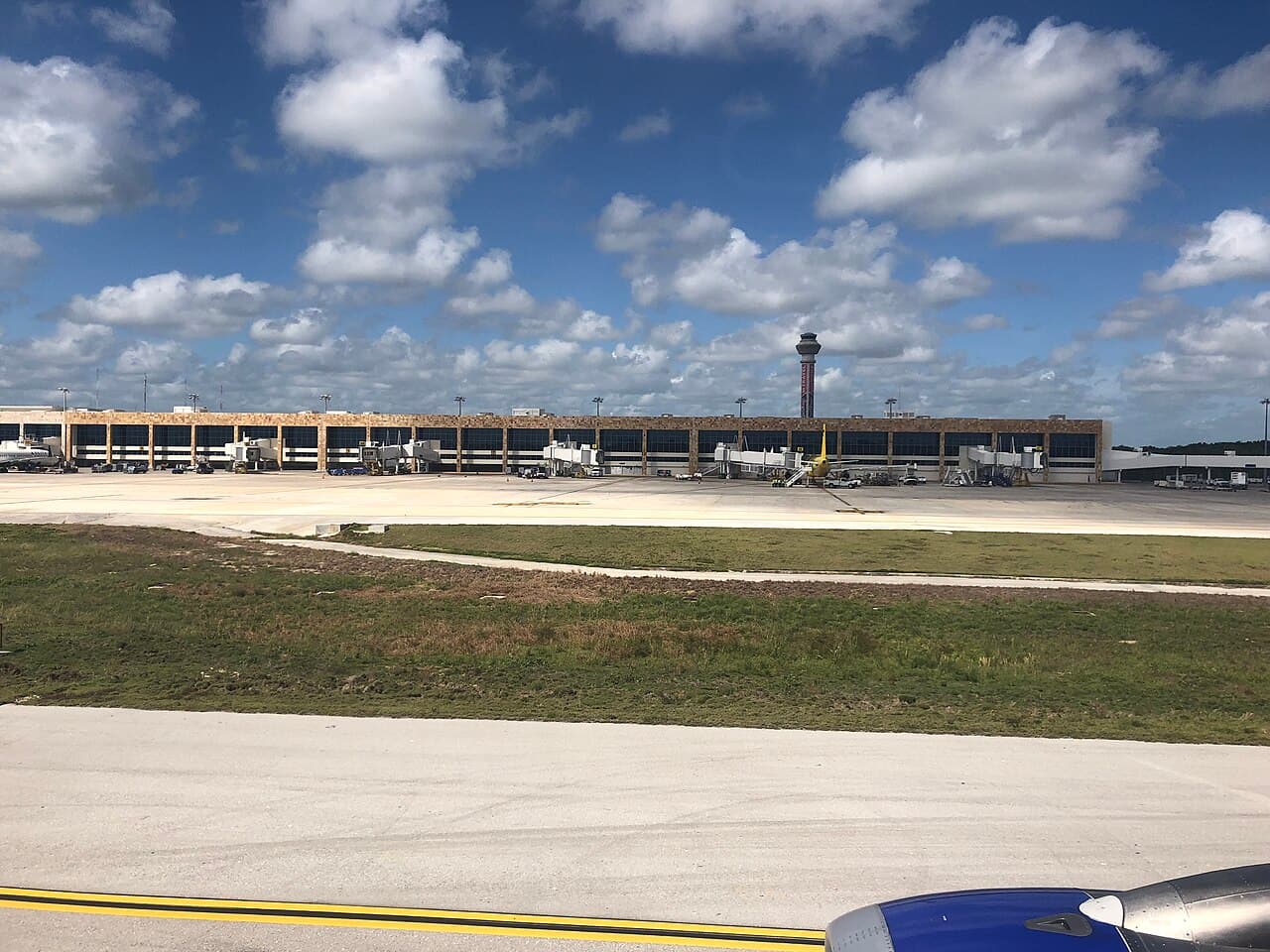 Vista desde una aeronave de la plataforma de un aeropuerto con terminal de amplios ventanales, puentes de abordaje, vehículos de servicio, una torre de control y cielo parcialmente nublado.