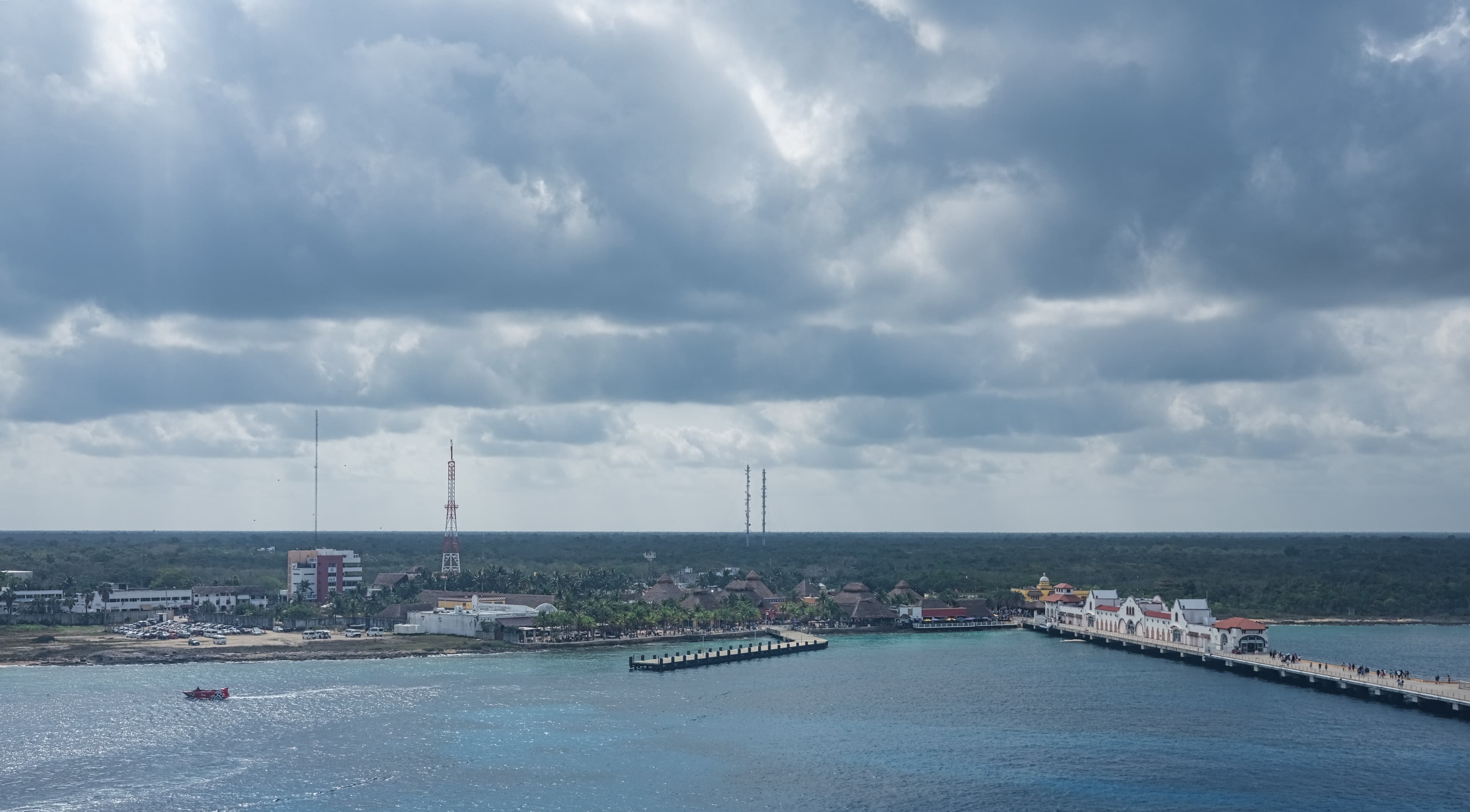 Vista panorámica del muelle y la costa de Mahahual, con el malecón de edificios blancos y techos rojos, palapas, torres de telecomunicaciones y lanchas bajo un cielo nublado.